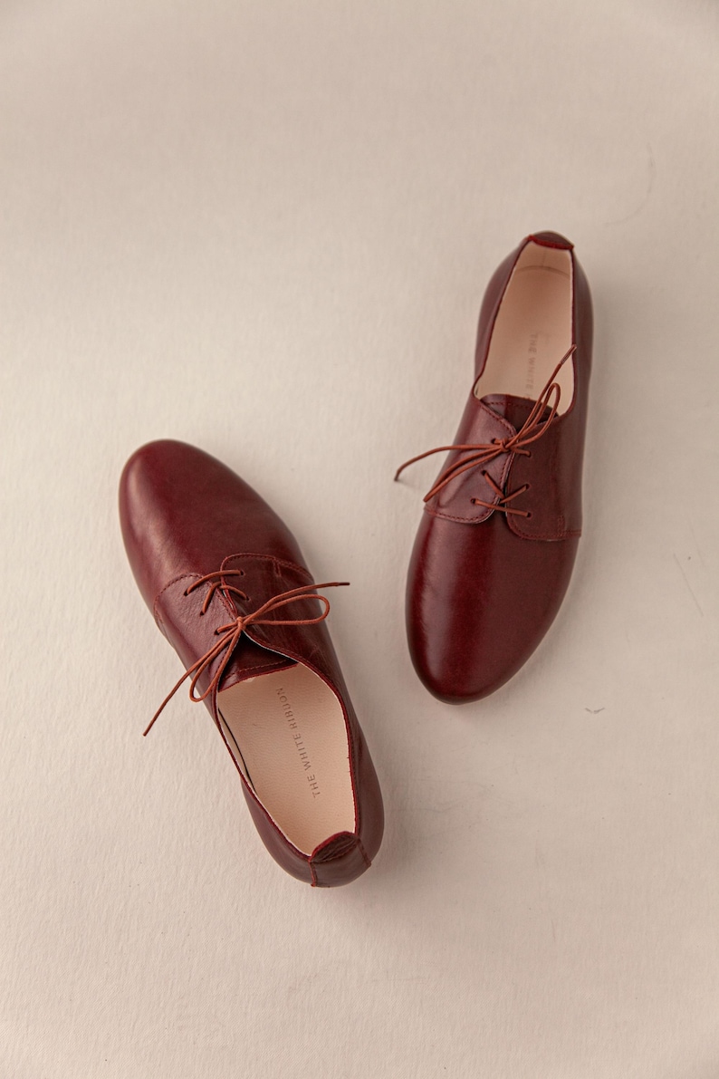 May include: A pair of burgundy leather Oxford shoes with matching laces. The shoes have a classic design with a rounded toe and a low heel. The interior of the shoe is visible, with the text "THE WHITE RIBBON" printed on the insole.