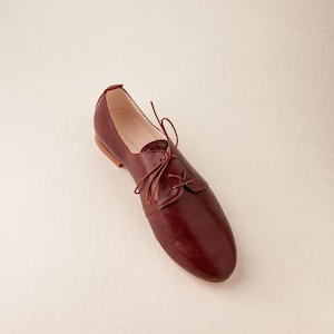 May include: A single burgundy leather oxford shoe with a lace-up closure. The shoe has a rounded toe and a low heel. The shoe is set against a plain, off-white background. The shoe is a size 9.