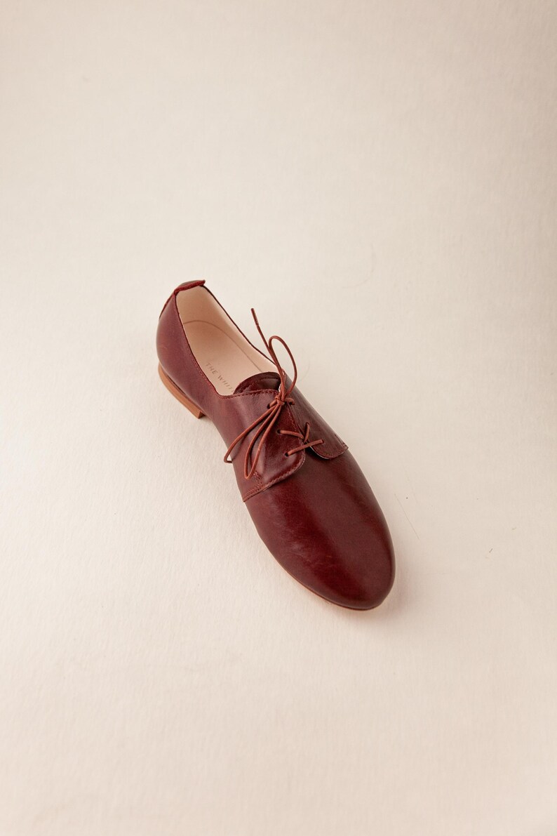 May include: A single burgundy leather oxford shoe with a lace-up closure. The shoe has a rounded toe and a low heel. The shoe is set against a plain, off-white background. The shoe is a size 9.