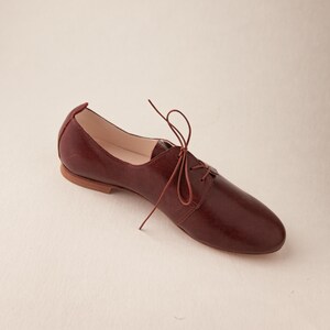 May include: A burgundy leather oxford shoe with a pointed toe and a lace-up closure. The shoe has a low heel and a smooth, polished finish. The laces are a matching burgundy color. The shoe is designed for everyday wear.