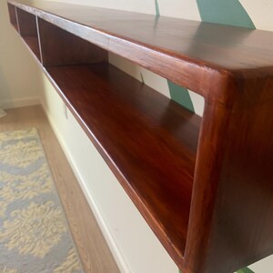 May include: A long, rectangular, floating shelf made of dark stained wood. The shelf has a smooth, glossy finish and features three open compartments. It is mounted on a white wall with a decorative painted design.