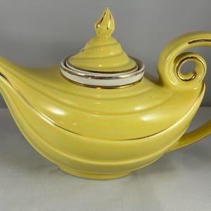 May include: A yellow ceramic teapot with a curved handle and spout. The lid has a spiral design and a gold finial. Gold trim accents the spout, handle, and lid. The teapot has a rounded, genie-lamp-like shape.