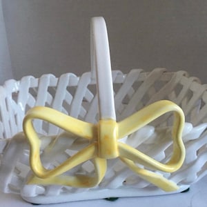 May include: White ceramic basket with a woven design and a curved handle. A yellow bow is attached to the front. The basket is rectangular and has a decorative appearance.