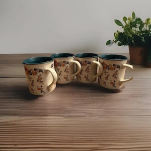 Può includere: Quattro tazze color crema con motivi floreali e bordi turchese sono disposte su una superficie di legno. Ogni tazza ha un manico curvo e un motivo floreale abbinato. Una pianta in vaso è visibile sullo sfondo.