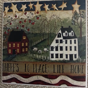 May include: A folk art painting depicts a rural scene with two houses, trees, and sheep. The artwork features a red house and a white house with a black roof. The text "THERE'S NO PLACE LIKE HOME" is at the bottom, with a red, white, and blue border.