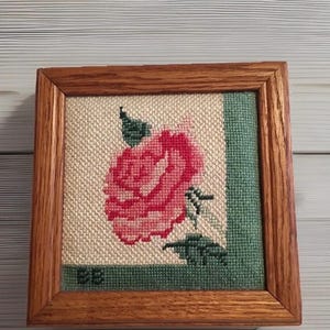 Może przedstawiać: Oprawiony haft krzyżykowy przedstawiający różową różę z zielonymi liśćmi i detalami. Dzieło sztuki jest osadzone na beżowym tle z zieloną obwódką i zamknięte w drewnianej ramie. Inicjały "BB" są wyszyte w lewym dolnym rogu.