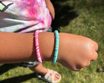 May include: Two colorful bracelets are worn on a person's arm. One bracelet is pink and white, and the other is blue and green. The bracelets are made of small, flat beads. The person is wearing a tie-dye shirt.