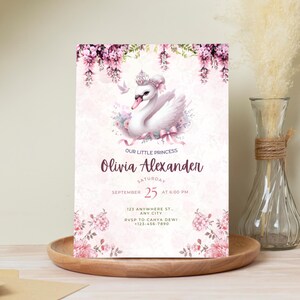 K&ouml;nnte beinhalten: Eine hellrosa Einladungskarte mit einer Schwan-Illustration mit Krone, umgeben von Blumen. Der Text lautet "Our Little Princess" und enth&auml;lt den Namen "Olivia Alexander", Datum, Uhrzeit und RSVP-Informationen. Die Karte liegt auf einem Holztablett.