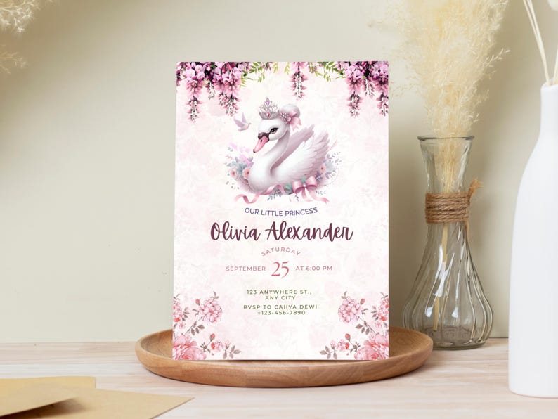 K&ouml;nnte beinhalten: Eine hellrosa Einladungskarte mit einer Schwan-Illustration mit Krone, umgeben von Blumen. Der Text lautet "Our Little Princess" und enth&auml;lt den Namen "Olivia Alexander", Datum, Uhrzeit und RSVP-Informationen. Die Karte liegt auf einem Holztablett.