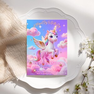K&ouml;nnte beinhalten: Eine farbenfrohe Einladungskarte mit einem Cartoon-Einhorn vor einem Himmel- und Wolkenhintergrund. Der Text lautet "Olivia WIRD 5" mit Veranstaltungsdetails. Die Karte liegt auf einem wei&szlig;en Teller.