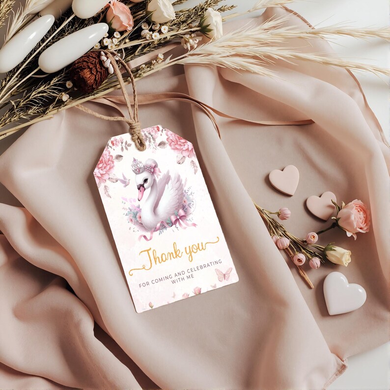 K&ouml;nnte beinhalten: Ein Geschenk-Anh&auml;nger mit einer Schwan-Illustration, floralen Akzenten und dem Text "Thank you". Der Anh&auml;nger ist mit Bindfaden befestigt und liegt auf einem weichen, rosa Stoff. Dekorative Elemente sind Herzen und Blumen.