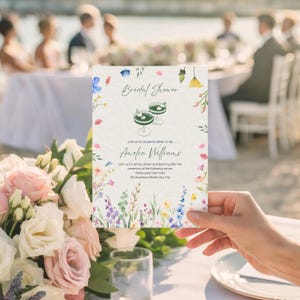 Könnte beinhalten: Eine florale Brautparty-Einladung mit dem Text "Bridal Shower" und "Amelia Williams". Die Einladung zeigt Aquarellblumen in Rosa, Blau und Gelb sowie zwei Cocktailgläser. Die Einladung wird vor einem verschwommenen Hintergrund gehalten.