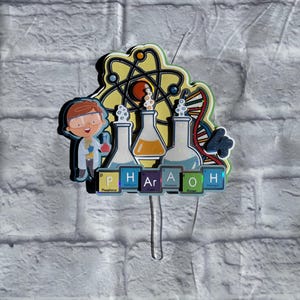 May include: A colorful cake topper featuring a cartoon scientist holding a beaker, with beakers, an atom, and DNA. The word "PHARAOH" is spelled out with periodic table elements. The topper is on a clear plastic stick.