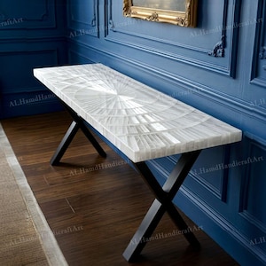 May include: A white console table with a geometric design on the top. The table has black, cross-shaped legs. It is set against a blue wall and a dark wood floor. The table's length is approximately 120 cm.