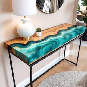 Puede incluir: Una consola moderna con una tapa de madera y un diseño de río de resina turquesa. La mesa tiene una estructura de metal negro y está colocada contra una pared neutra. Una lámpara blanca y una pequeña planta están sobre la mesa.