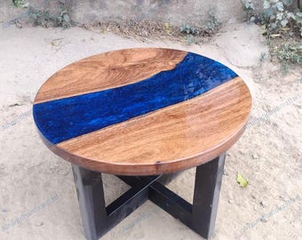 May include: Round wooden side table with a blue resin river design. The tabletop is made of natural wood with a glossy finish, and the blue resin flows through the center. The table has a black, X-shaped base.
