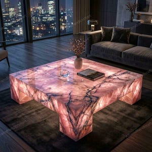 Puede incluir: Una mesa de centro cuadrada hecha de piedra rosa translúcida con vetas oscuras prominentes. La mesa cuenta con un sistema de iluminación interno que emite un brillo suave a través de la superficie pulida, la cual sostiene un jarrón y un libro.