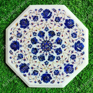 Puede incluir: Tablero de mesa octogonal de mármol blanco con un diseño floral incrustado. El diseño presenta flores azules y detalles dorados. El tablero de la mesa está sobre un fondo verde. El texto "ALHamdHandicraftArt" es visible.
