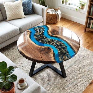 May include: A round coffee table featuring a natural wood slab with a live edge and a central river design made of translucent blue resin filled with smooth, multi-colored river stones. The table is supported by a black metal frame base.