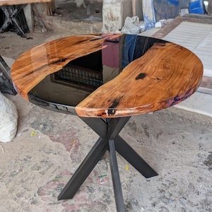 May include: A round table featuring a natural wood slab top with a glossy black resin centre inlay. The wood displays prominent grain patterns and organic edges. The table is supported by a black metal base with four intersecting legs.