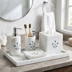 Può includere: Un set di accessori da bagno in marmo bianco composto da un dispenser per sapone, un portaspazzolino, un portasapone e un copri scatola per fazzoletti su un vassoio rettangolare. Ogni pezzo è decorato con un piccolo motivo floreale intarsiato.