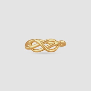 Puede incluir: Un anillo dorado con un diseño de nudo. El anillo está hecho de un material metálico brillante y se muestra sobre un fondo gris claro. El diseño del nudo es el punto focal del anillo.