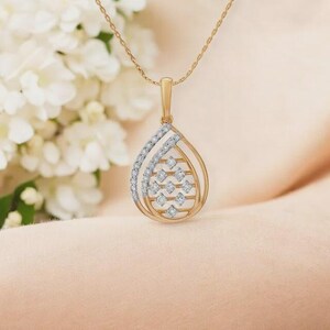 Pode incluir: Um colar com pingente de ouro com um design em forma de gota. O pingente apresenta pequenos diamantes nas bordas e dentro da forma de gota. O colar é exibido em um fundo macio e neutro.