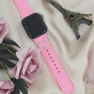 May include: A pink smartwatch band with a black watch face. The band features a silver charm with a heart and infinity symbol design, adorned with small, sparkling crystals. A miniature Eiffel Tower and pink roses are in the background.