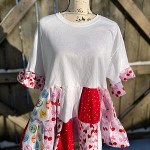May include: White t-shirt with a tiered, patchwork skirt. The skirt features red, pink, and white fabrics with heart, cherry, and candy-themed patterns. The sleeves have ruffled edges with cherry prints. The shirt is displayed on a mannequin.