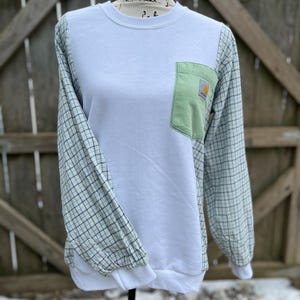 May include: White sweatshirt with green plaid sleeves and a light green pocket. The pocket features a Carhartt logo. The sleeves and pocket are a different material than the white body of the sweatshirt. The sweatshirt is displayed on a mannequin.
