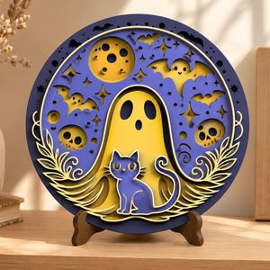 May include: A layered, circular Halloween decoration featuring a yellow ghost, a blue cat, bats, skulls, and a moon against a purple background. The design is set in a dark blue frame and displayed on a wooden stand, creating a 3D effect.