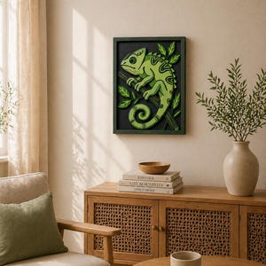 May include: A framed, layered wood art piece featuring a green chameleon perched on a branch. The artwork has a dark green frame and a black background. The chameleon is depicted in shades of green with detailed leaf accents, creating a nature-inspired wall decor.