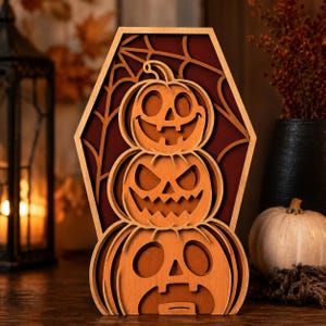 Peut inclure: Une décoration en bois superposé en forme de cercueil présentant trois citrouilles d'Halloween empilées avec des expressions faciales sculptées. Le fond à l'intérieur du cadre affiche une surface rouge foncé avec une toile d'araignée découpée.
