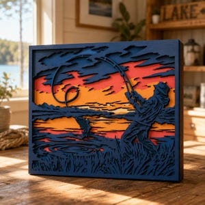 Pode incluir: Uma peça de arte de parede retangular de madeira em várias camadas com a silhueta de uma pessoa pescando ao pôr do sol. O design inclui camadas azul-escuras para o primeiro plano e o céu, com camadas vibrantes de laranja e vermelho.