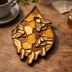 Può includere: Sottobicchiere in legno a forma di goccia, con un design a nido d'ape e tre api. Il sottobicchiere è realizzato in legno a strati nei toni del marrone e del beige. Il design include un effetto miele gocciolante.