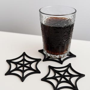 Pode incluir: Um copo transparente com uma bebida escura repousa sobre um porta-copos preto em forma de teia de aranha. Dois porta-copos pretos adicionais em forma de teia de aranha são visíveis. O copo tem um padrão texturizado. Os porta-copos são de material sólido.