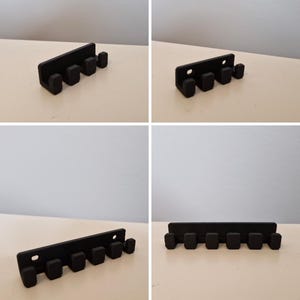 May include: A black wall-mounted key rack with six pegs. The rack is rectangular with rounded edges and two holes for mounting. The pegs are evenly spaced and cylindrical. The key rack is made of a solid, matte black material.