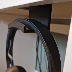 May include: Black headphones hanging from a black under-desk headphone mount. The headphones have a dark gray headband and earcups. The mount is attached to the underside of a white shelf.