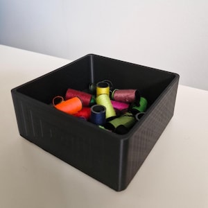 May include: A black square container filled with colorful spools of thread. The thread colors include orange, red, yellow, green, blue, and pink. The container is sitting on a white surface.