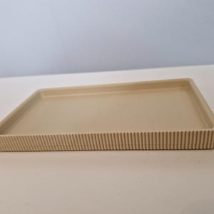 May include: A rectangular beige tray with a ribbed edge. The tray has a simple, minimalist design and is made of a solid, neutral-toned material. The ribbed detailing adds a subtle texture to the tray's edge.