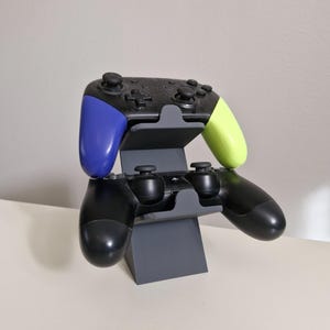 Puede incluir: Un soporte gris impreso en 3D que sostiene dos mandos de juego. El mando superior tiene detalles en azul y verde. El mando inferior es negro. El soporte está diseñado para sujetar los mandos en posición vertical.