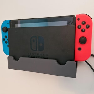 Puede incluir: Una consola Nintendo Switch acoplada en un soporte de pared gris. La consola tiene una pantalla negra y una base negra con el logotipo de Nintendo Switch. Los mandos Joy-Con son azules y rojos. Un cable negro está conectado.