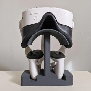 Op de afbeelding: Een witte en zwarte VR-headset en controllers worden weergegeven op een grijze standaard. De standaard is ontworpen om de headset en twee controllers te bevatten. De VR-headset heeft een zwart gezichtskussen en een witte buitenkant. De controllers zijn wit met zwarte bovenkanten.