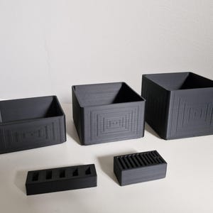 May include: A collection of dark gray storage containers and organizers. The set includes three square boxes of varying sizes and two rectangular organizers. Each piece features a geometric design on the exterior, adding a modern touch to the functional items.