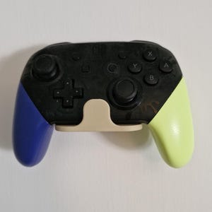 Puede incluir: Un mando de videojuegos negro y multicolor con agarres azules y verdes. El mando tiene una cruceta, palancas analógicas y botones de acción. Está montado en un soporte beige.