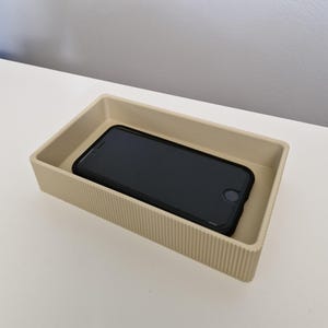 Może przedstawiać: Beżowa prostokątna taca z pionowymi żebrowanymi bokami mieści czarny smartfon. Taca wykonana jest z solidnego materiału i stoi na białej powierzchni. Smartfon ma czarne etui.