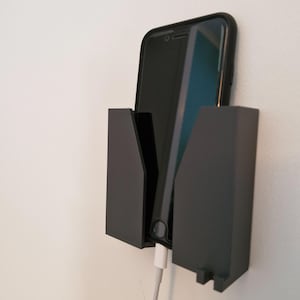 May include: A black phone is secured in a black wall-mounted holder. The phone is charging with a white cable. The holder has a minimalist design and is mounted on a white wall. The phone case is black.