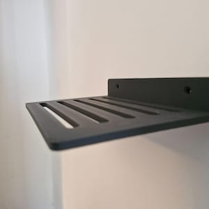 Puede incluir: Una estantería de pared negra mate con un diseño ranurado. La estantería tiene forma rectangular con una superficie plana y una parte trasera elevada, con dos orificios pretaladrados para el montaje. La estantería está diseñada para exhibir objetos.