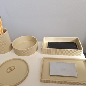 May include: A set of beige desk tidies, including a pencil pot with pencils, an oval dish, a rectangular tray with a phone, and a square tray with a small laptop. The items have a ribbed texture.