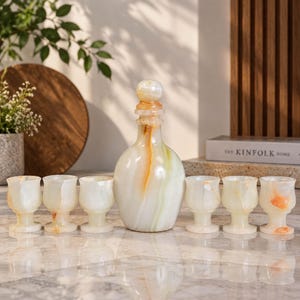 Può includere: Un set di sei piccoli calici e una caraffa abbinata con tappo, realizzati in pietra lucida con venature traslucide bianche, color crema e arancio-marrone. La caraffa presenta un corpo arrotondato e un collo stretto.